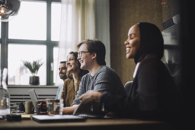 Cheerful young businessman sitting by male and female colleagues while discussing in meeting room