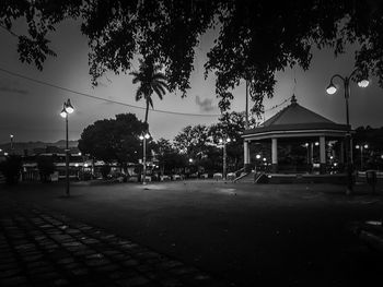 Illuminated street by building against sky at dusk