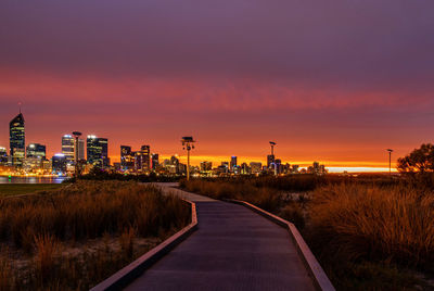 Road by illuminated buildings against sky during sunset