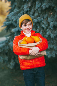 A boy in a bright orange jacket and a yellow hat holds a large package with oranges in his hands 