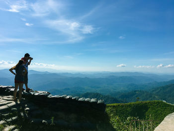 Woman standing by man photographing on cliff against mountains