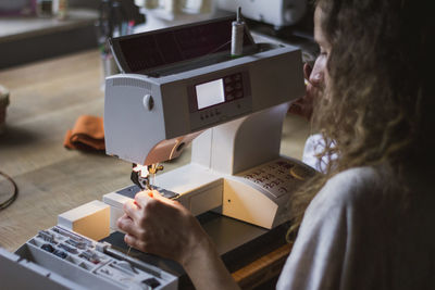 Back view of anonymous crop woman using sewing machine making clothes sitting at table in house