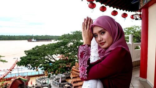 Portrait of young woman standing by railing against sky