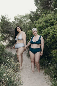 Positive curvy female friends in swimwear walking on sandy path on beach and enjoying summer looking at camera