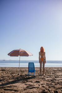 Back view of anonymous young redhead woman standing on beach on a sunny day in summer