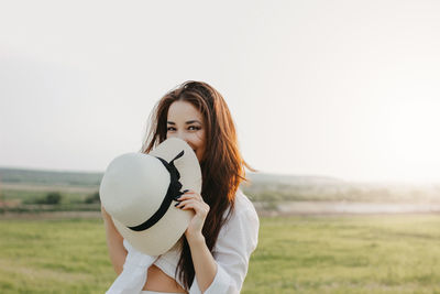 Portrait of young woman holding hat on field against sky