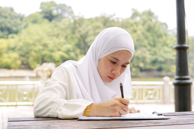 Close-up of young woman wearing hijab writing in paper on table against trees