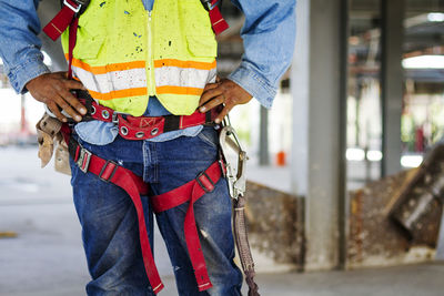 Midsection of manual worker wearing safety harness standing at construction site