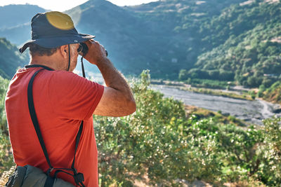 Rear view of tourist man looking through binoculars at the mountains view. hiking, unity with nature