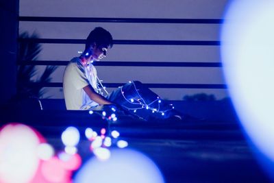 Young man with illuminated fairy lights while sitting outdoors at night