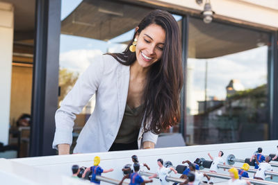 Happy adult hispanic woman in smart casual clothes smiling and playing table football during party on weekend day on terrace