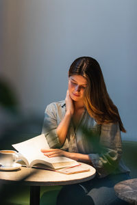 Young serene female touching face while reading textbook at cafeteria table with cup of hot drink