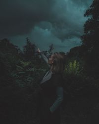 Young woman with hand raised standing amidst trees in forest against cloudy sky at dusk