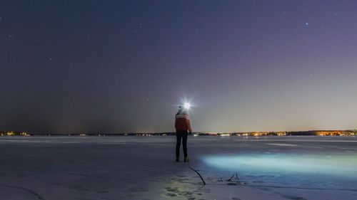 Rear view full length of person standing on snow covered landscape against sky at night
