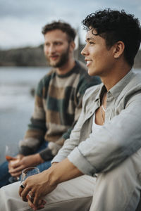 Young man enjoying wine with male friend sitting near lake