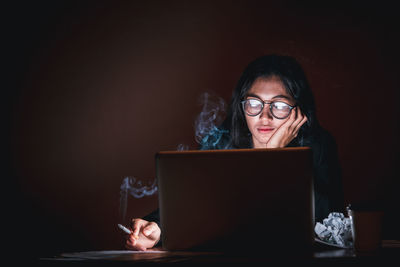 Depressed woman smoking cigarette while using laptop in darkroom