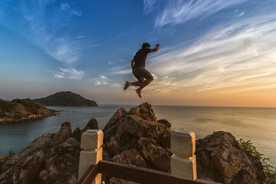 Man jumping on rock by sea against sky