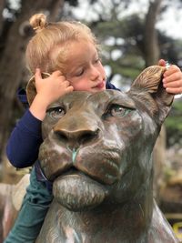 Close-up of girl sitting on lioness statue against trees