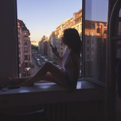 Side view of woman using phone while sitting on window sill