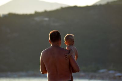 Rear view of shirtless father holding son while standing outdoors