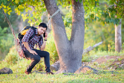 Smiling man sitting on hammock while talking on smart phone in forest