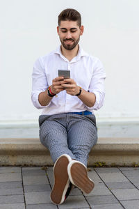 Young bearded man sitting against a white wall using the mobile phone