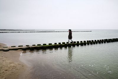 Women standing on wooden post in sea against clear sky