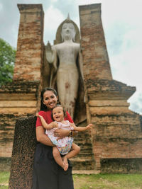 Woman with baby stands in front of stone buddha figure in buddhist temple