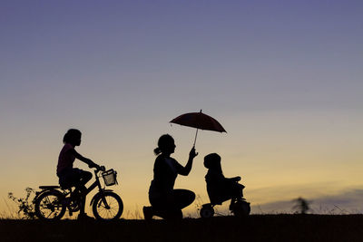 Silhouette woman with daughters riding bicycles on field against sky during sunset