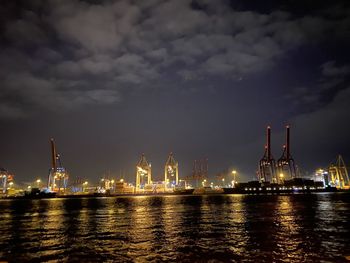 Illuminated factory by sea against sky at night