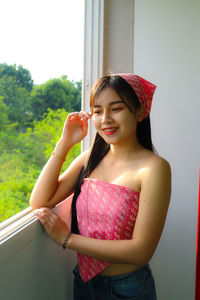 Portrait of smiling young woman standing against window