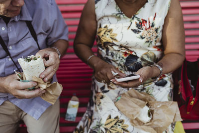 Senior man with food sitting by woman using smart phone on bench