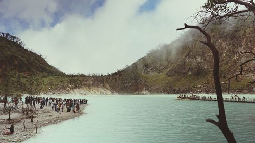 People at lakeshore with mountains in background against cloudy sky