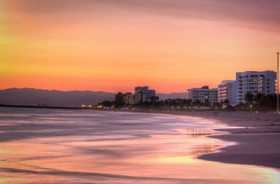 Scenic view of beach by buildings against orange sky during sunset