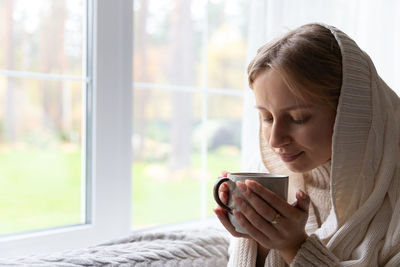 Woman drinking hot tea while resting at home in morning, sitting by window. autumn time, flu season.