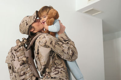 Side view of happy female soldier in uniform with backpack holding kissing cute son in bedroom