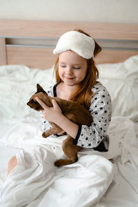 A little girl with red hair sits in the bedroom on a bed with white sheets and hugs her ginger cat. 
