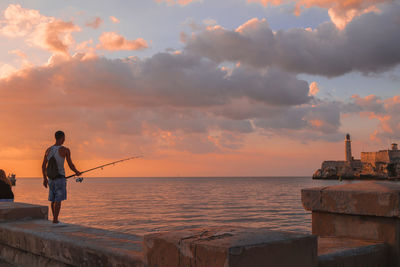 Rear view of man fishing in sea against sunset sky