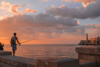 Rear view of man fishing in sea against sunset sky