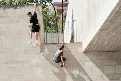 Back view of brave male friends hanging on wall of concrete building while performing risky trick and doing parkour in city