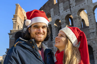 A young couple poses in front of the colosseum in red santa claus hats.