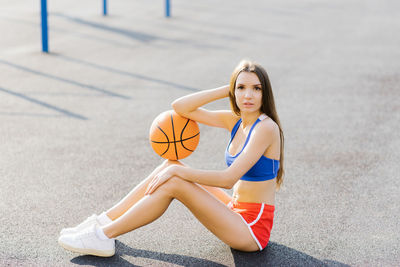 A young athletic woman sits on the floor and holds a basketball in a sports stadium, outdoors