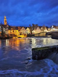 Illuminated buildings by river against sky at dusk