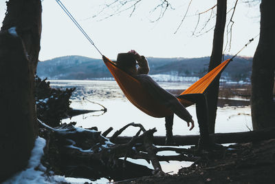 Full length of woman wearing hat resting in hammock by lake against sky