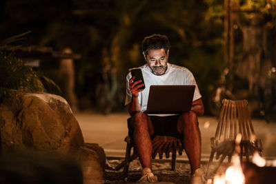 Smiling man with phone using laptop at night