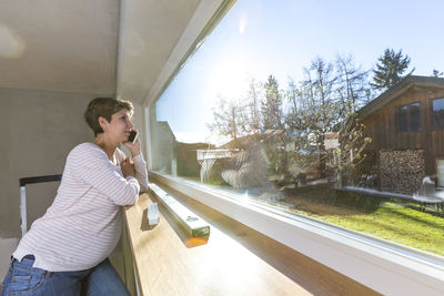 Woman talking on mobile phone while looking through window at home