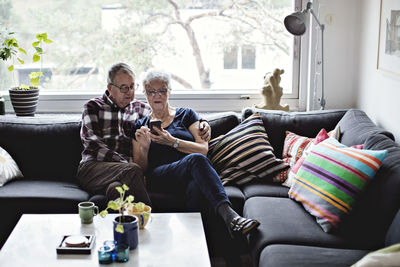 Senior couple sharing smart phone sitting on sofa in living room against window at home