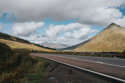 Empty road by mountains against cloudy sky