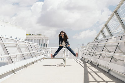 Full length of playful woman cycling on bridge in city during sunny day