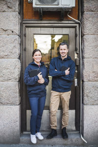 Full length portrait of smiling engineers holding computer equipment while standing outside electronics store